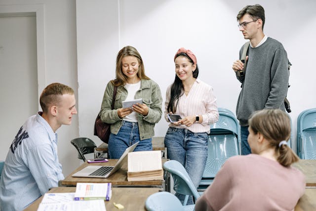 A group of five people are gathered in a room, smiling and engaged in conversation. Two are seated at a table with laptops and papers, while three stand nearby, holding notebooks.