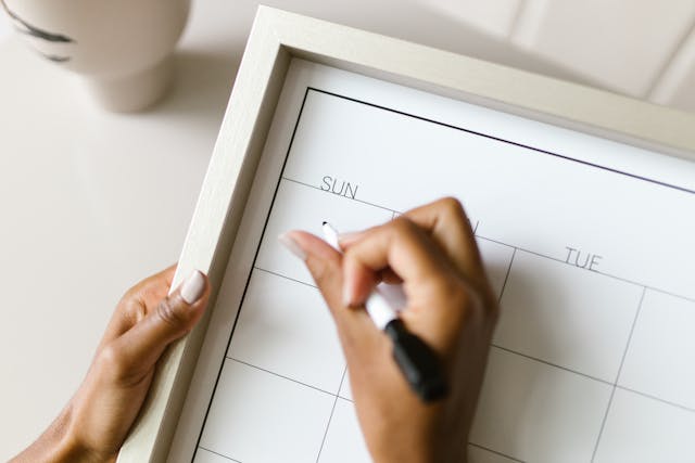A person writes on a blank calendar board on a desk. The board shows days of the week starting with Sunday.